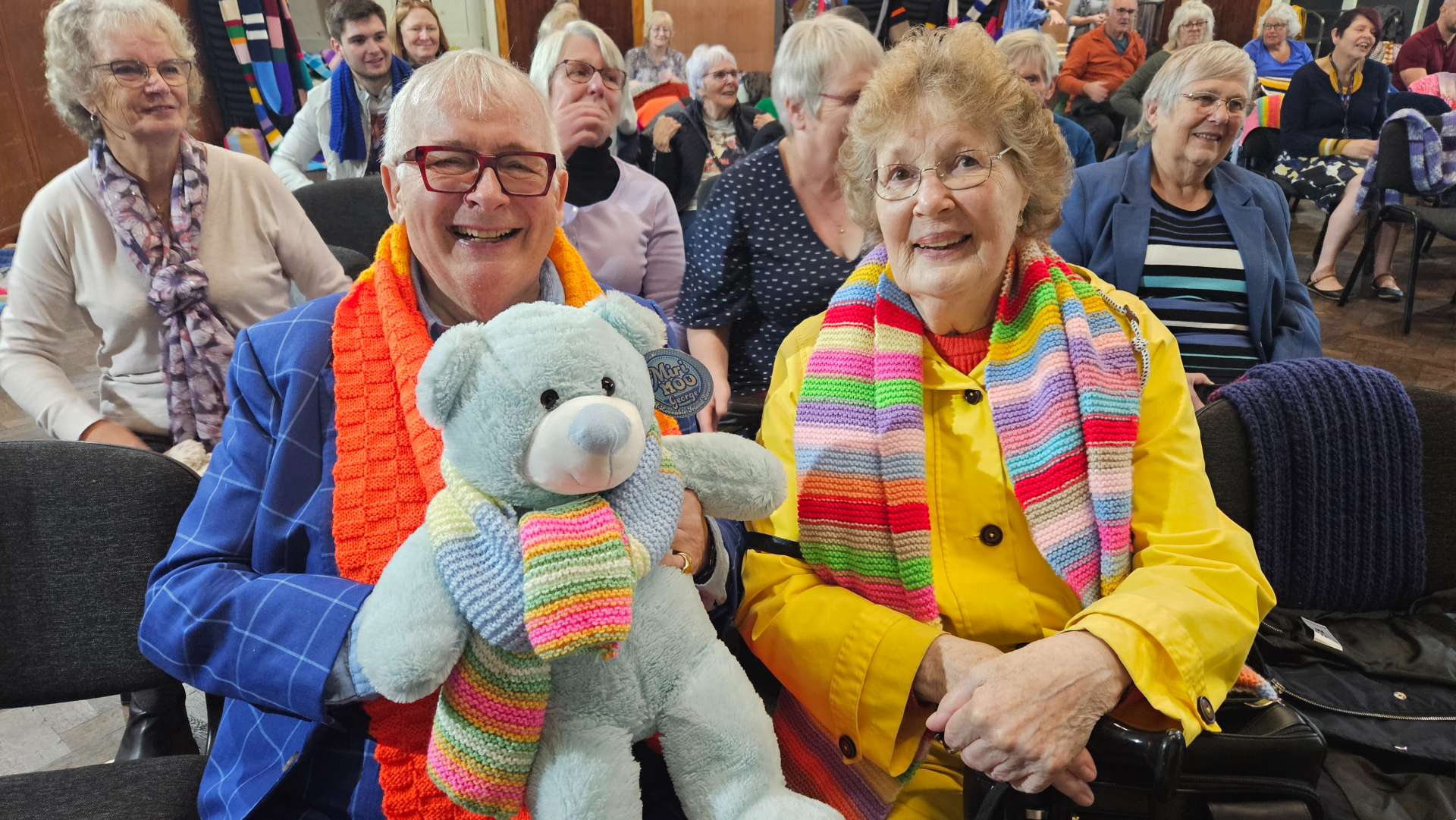 Christopher Biggins holding a blue teddy bear with scarves and surrounded by women also in scarves at Scarf For Lewy event